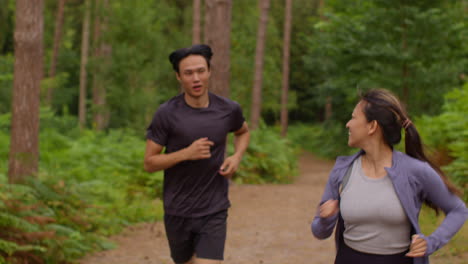 Mid-Adult-Couple-Exercising-Doing-Work-Out-Outdoors-Running-Through-Forest-With-Woman-Encouraging-Tired-Man-Wearing-Sports-Clothing-1